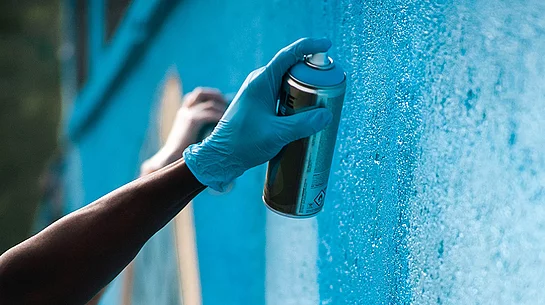 Eine Hand hält eine Sprühdose und sprayt ein Graffiti an eine Wand.